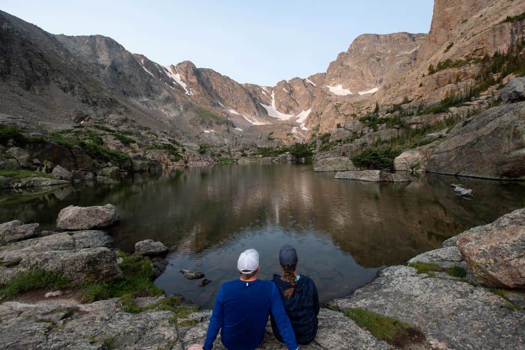 2 hikers sit in the center of the frame at the bottom, admiring Lake of Glass and the rocky peaks in the backdrop.