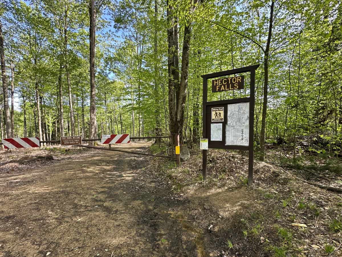 Trailhead entrance to Hector Falls in the Allegheny National Forest, showing a dirt road blocked by a gate and a wooden sign reading “HECTOR FALLS.” Forest trees line both sides of the entrance, with trail maps and hiking information posted on the signboard.
