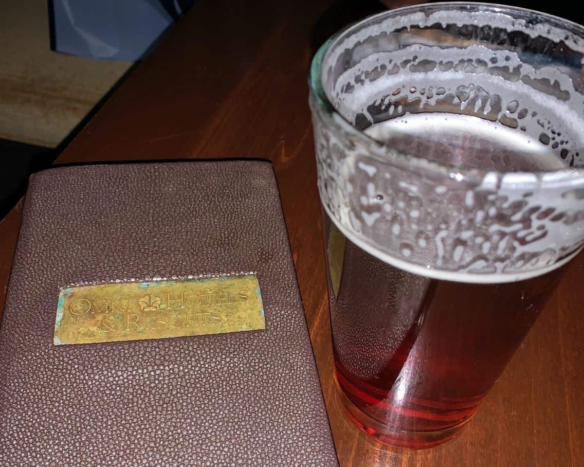 Close-up of an Omni Hotels & Resorts leather menu cover beside a half-finished pint of amber beer, representing the relaxed tavern dining at Omni Bedford Springs Resort.