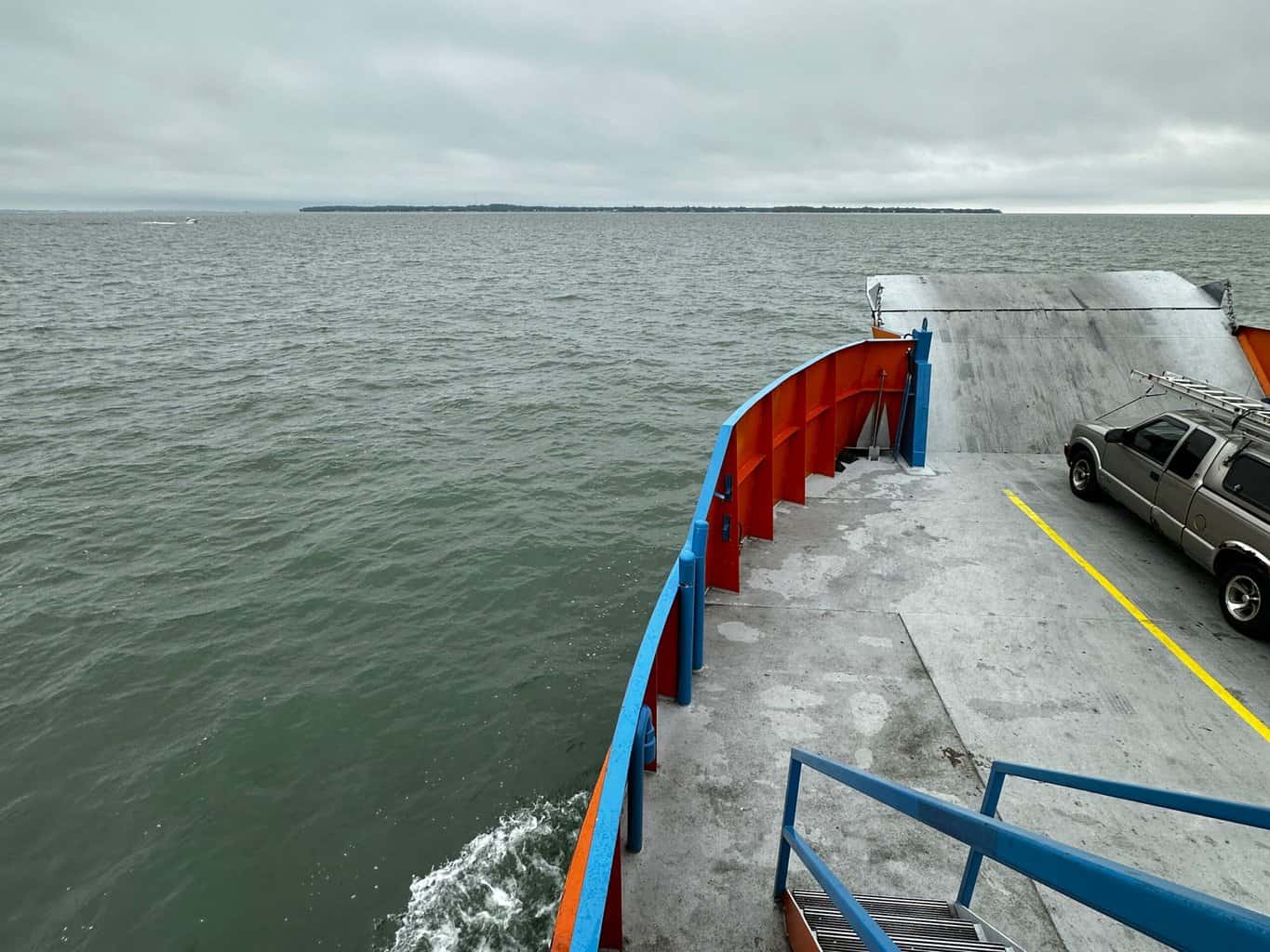 View from a ferry deck approaching Kelleys Island on a cloudy day, with a gray truck parked near the front and choppy water in the foreground.