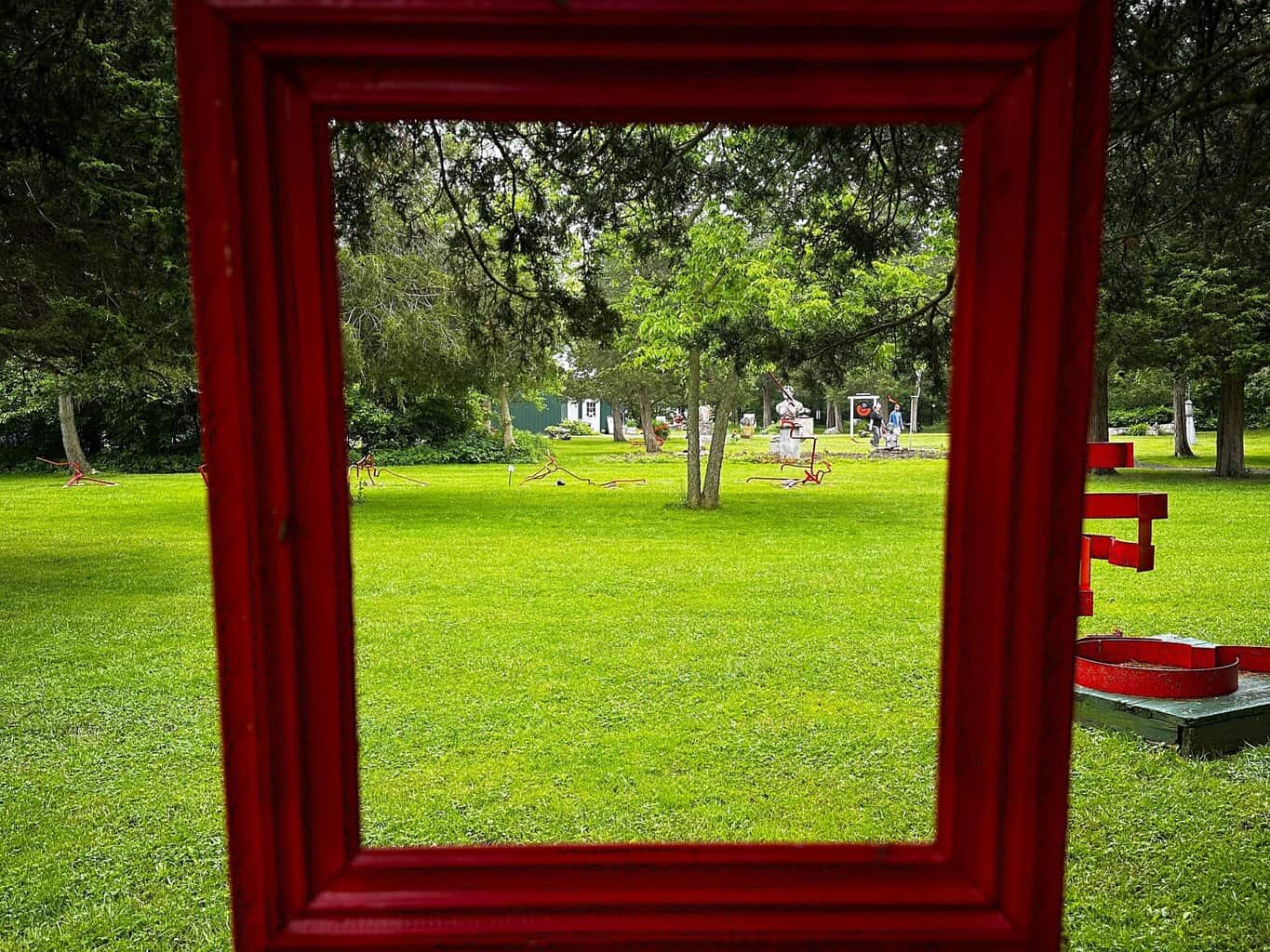 Sculptures and trees on a grassy lawn seen through a large red wooden frame, part of an outdoor art display on Kelleys Island.