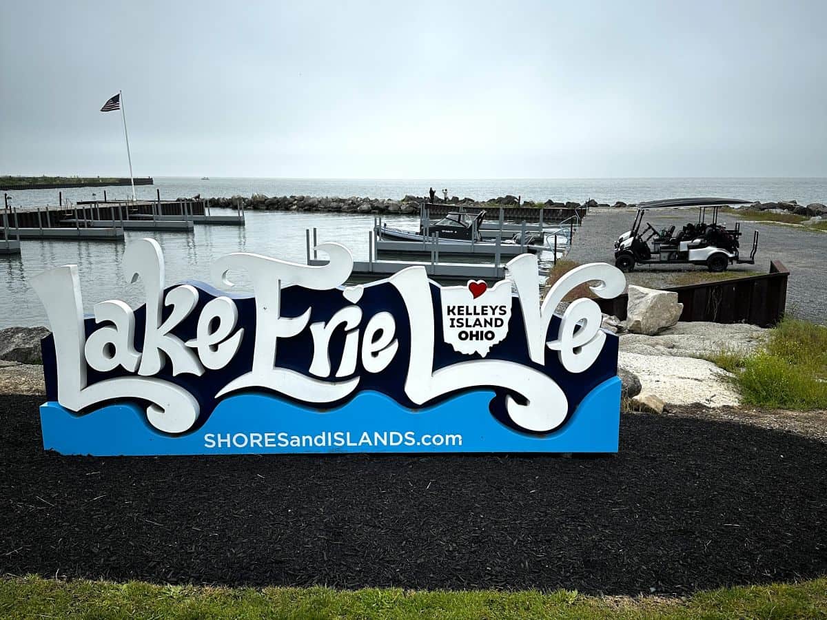 Decorative “Lake Erie Love” sign with a heart-shaped cutout reading “Kelleys Island Ohio” near a quiet marina, with golf carts parked by the water and the lake in the background.
