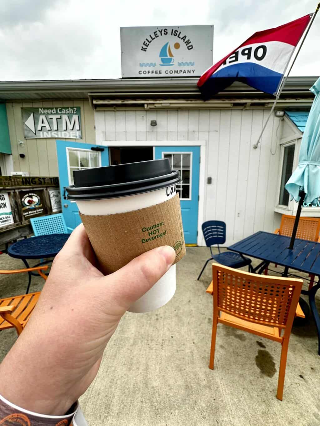 Hand holding a takeaway coffee cup in front of Kelleys Island Coffee Company, a small café with colorful outdoor seating and an open sign waving.