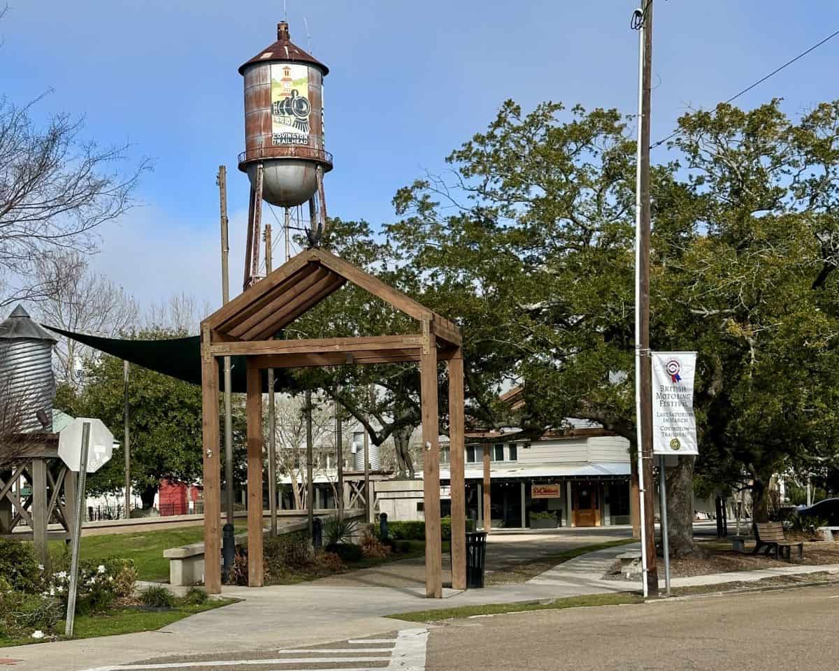 Historic water tower painted with Covington Trailhead artwork rises above a wooden pavilion entrance along the Tammany Trace in Covington Louisiana. The landmark marks a popular starting point for biking the Tammany Trace on the Louisiana Northshore.