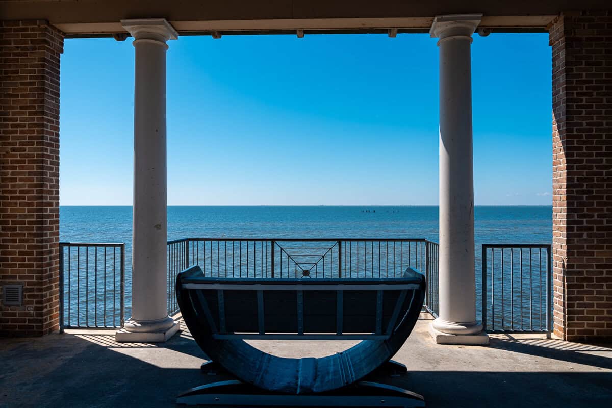 A historic covered brick pier with tall white columns frames a view of Lake Pontchartrain beyond a metal railing. A wooden boat bench sits in the center overlooking the water at Fontainebleau State Park, a great spot to take a break on a Northshore bike ride.