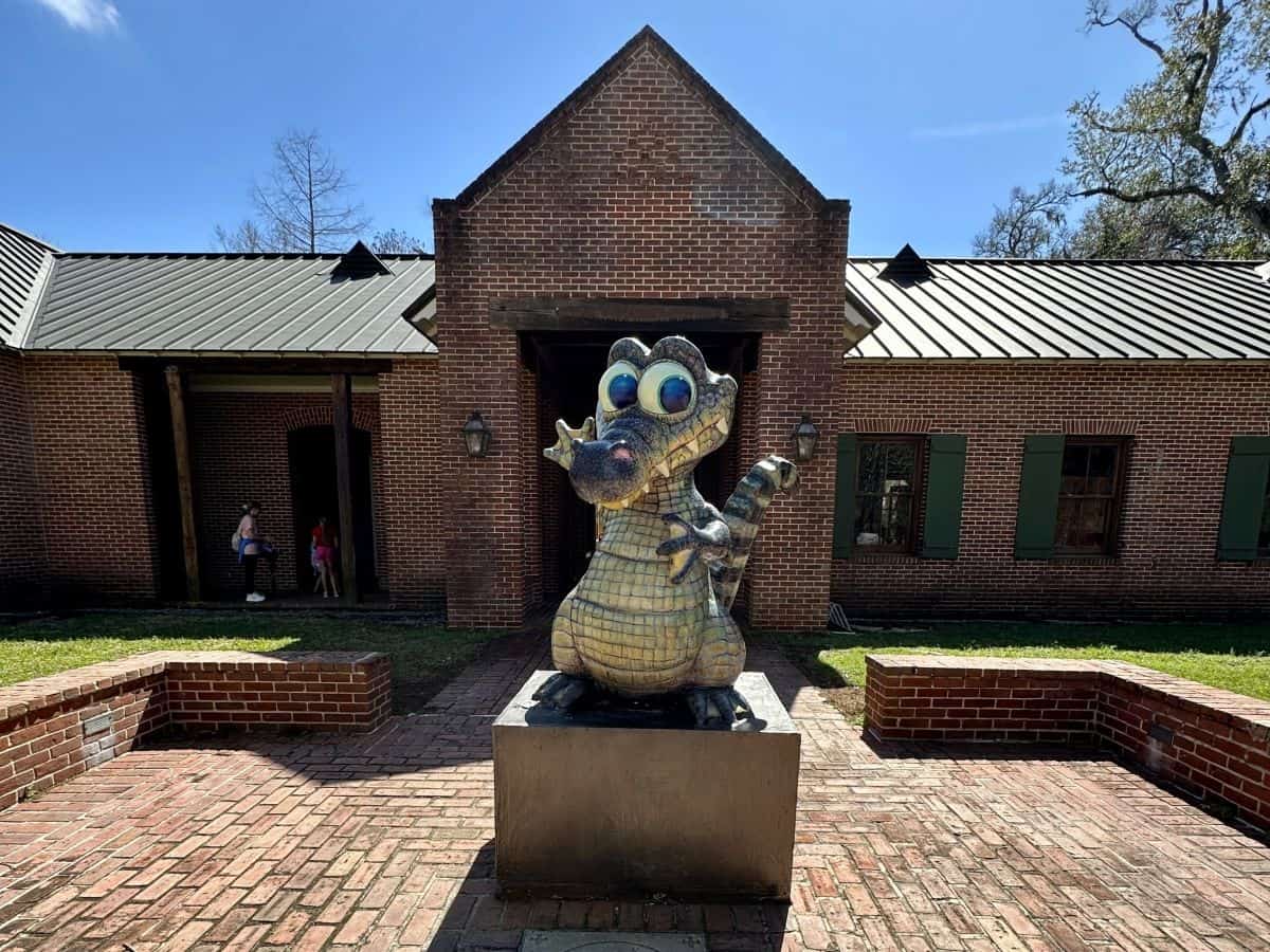 A playful cartoon alligator statue with big blue eyes stands on a pedestal in front of a historic brick building at Fontainebleau State Park. The colorful sculpture welcomes visitors to the park along Louisiana’s Northshore.