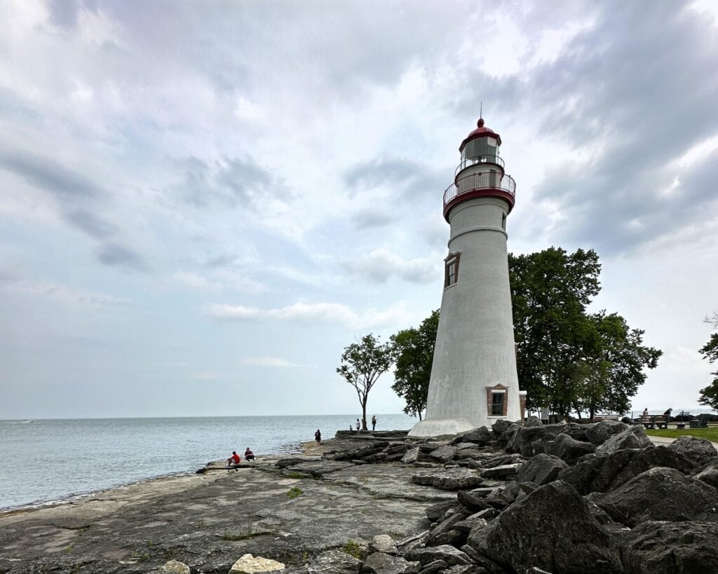 White lighthouse with a red lantern stands on a rocky shoreline along Lake Erie under a cloudy sky. Visitors walk and sit along the water near the historic Marblehead Lighthouse in Ohio.