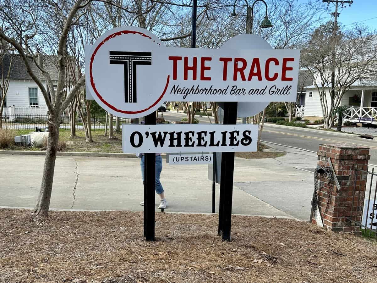 Sign reading O’Wheelies upstairs beneath The Trace Neighborhood Bar and Grill logo along a quiet street lined with trees and houses. The sign marks a popular stop near the Tammany Trace for food and drinks and where we rented our e-bikes during our day exploring the Northshore by bike.