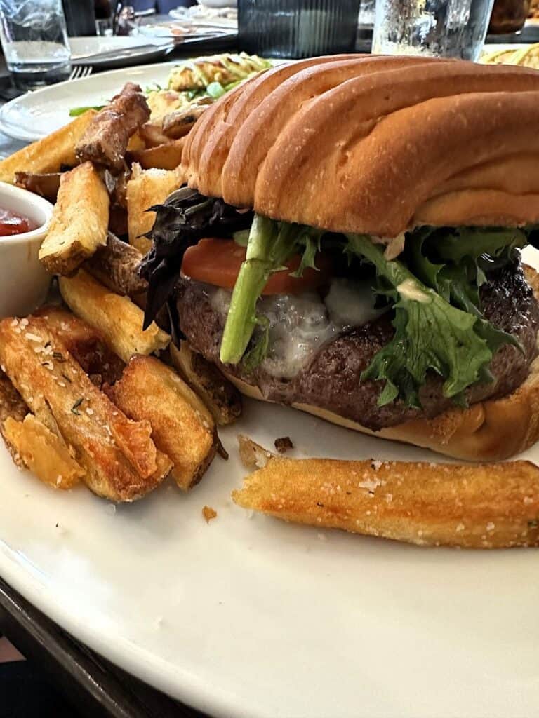 Cheeseburger with melted cheese, lettuce, and tomato on a toasted bun served with thick cut fries and ketchup. The burger is plated on a white dish at a restaurant table at Beach Glass Lounge in Marblehead, Ohio.