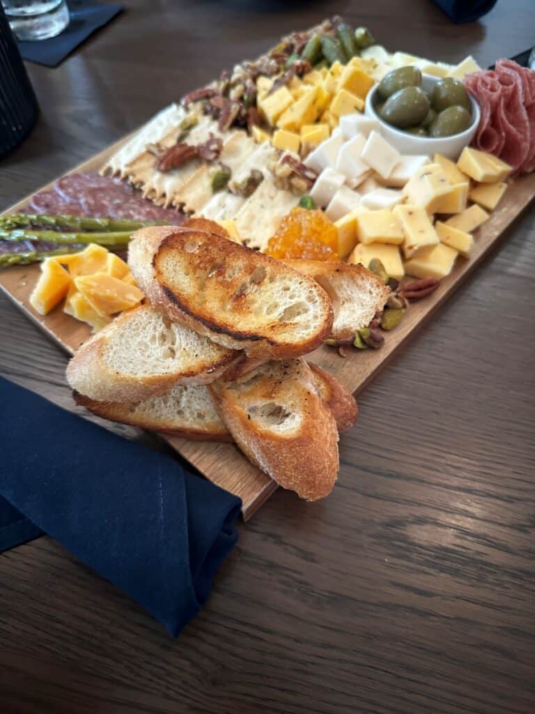 Wooden charcuterie board topped with sliced cheeses, cured meats, olives, nuts, crackers, pickles, and toasted crostini at Beach Glass Lounge in Marblehead, Ohio. The shareable appetizer features a mix of savory snacks and spreads arranged on a restaurant table.