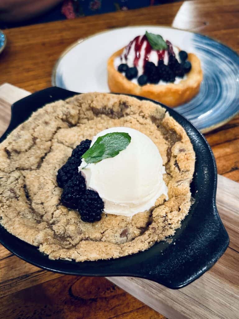 Warm skillet chocolate chip cookie topped with a scoop of vanilla ice cream, fresh blackberries, and mint. A small butter cake with whipped cream and berry sauce sits on a plate in the background at the Wave Lakeside Marblehead, Ohio.