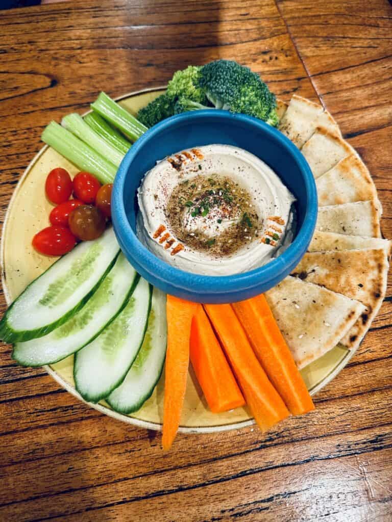 Colorful hummus platter surrounded by cucumber slices, carrot sticks, cherry tomatoes, celery, broccoli, and toasted pita wedges. The hummus is topped with olive oil and spices in a blue bowl at the center from the Wave Lakeside Marblehead, Ohio.