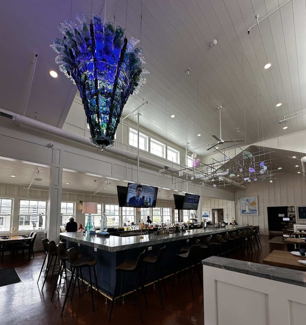 Bright restaurant interior with a long bar, large blue glass chandelier, and modern hanging art installation above the seating area at Beach Glass Lounge near Marblehead, Ohio. The airy dining room features tall windows and a coastal inspired design.