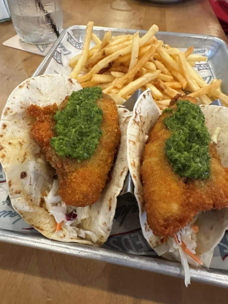 Two crispy fish tacos topped with green sauce and cabbage slaw served with a side of thin fries. The tacos sit in metal holders on a tray lined with paper at Red's Lakeside in Marblehead, Ohio.