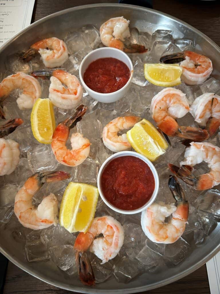 Shrimp cocktail arranged over crushed ice with lemon wedges and two cups of cocktail sauce. The chilled shrimp are spread across a large metal serving tray at Beach Glass Lounge in Marblehead, Ohio.