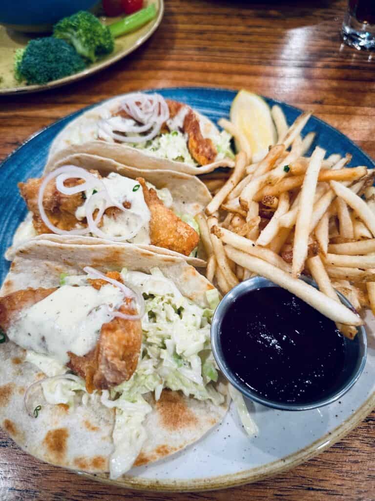 Plate of crispy fish tacos topped with cabbage slaw and creamy sauce served with fries and a side of dipping sauce. The casual seafood dish is arranged on a blue ceramic plate at the Wave Lakeside Marblehead, Ohio.