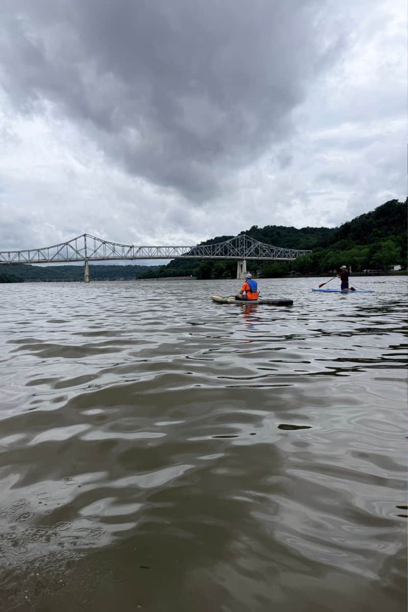 Two people paddleboard on the Kanawha River near a steel truss bridge in Winfield, West Virginia, beneath dark storm clouds.