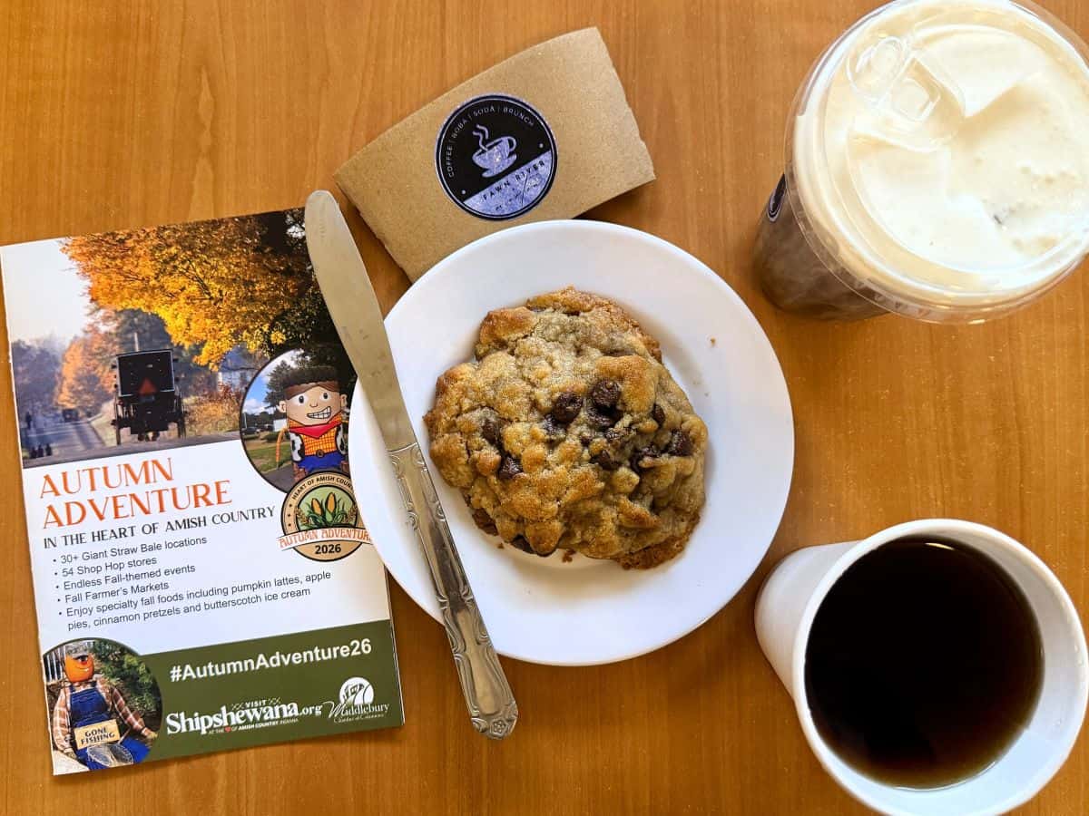 A chocolate chip cookie, an iced coffee, and a cup of black coffee sit beside a Shipshewana Autumn Adventure brochure on a wooden table at Fawn River + Morton.