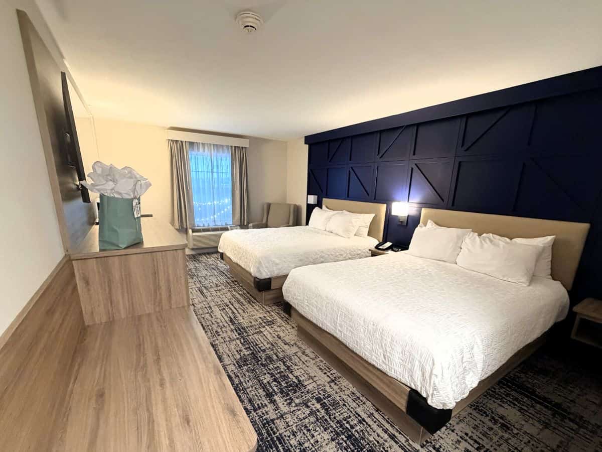 Renovated guest room at Blue Gate Garden Inn in Shipshewana, with two white beds against a dark blue accent wall, a desk, and a chair by the window