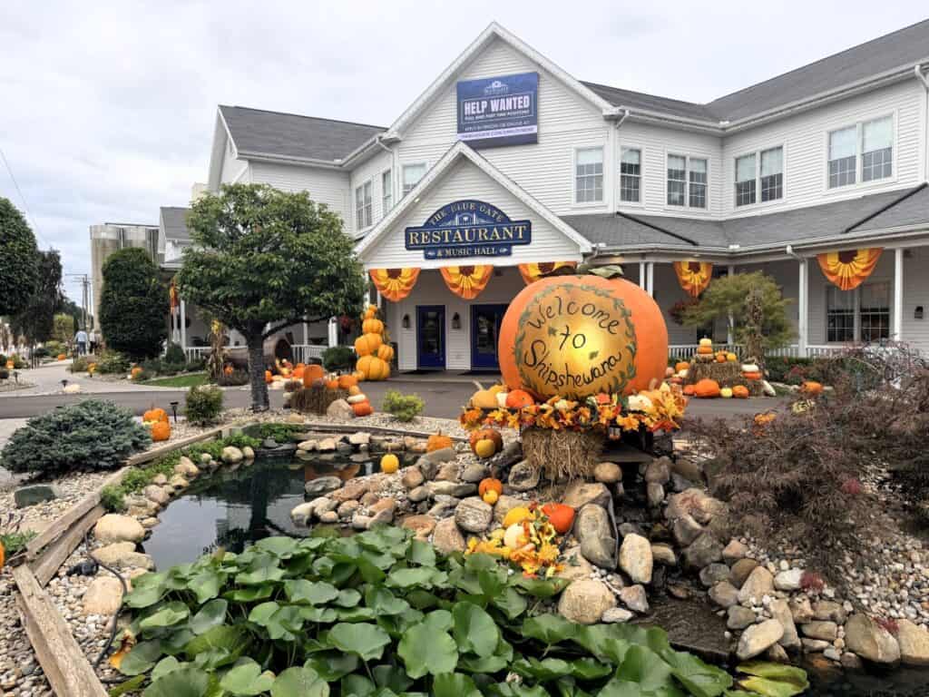 A giant “Welcome to Shipshewana” pumpkin and fall decorations outside Blue Gate Restaurant and Music Hall, beside a small landscaped pond.