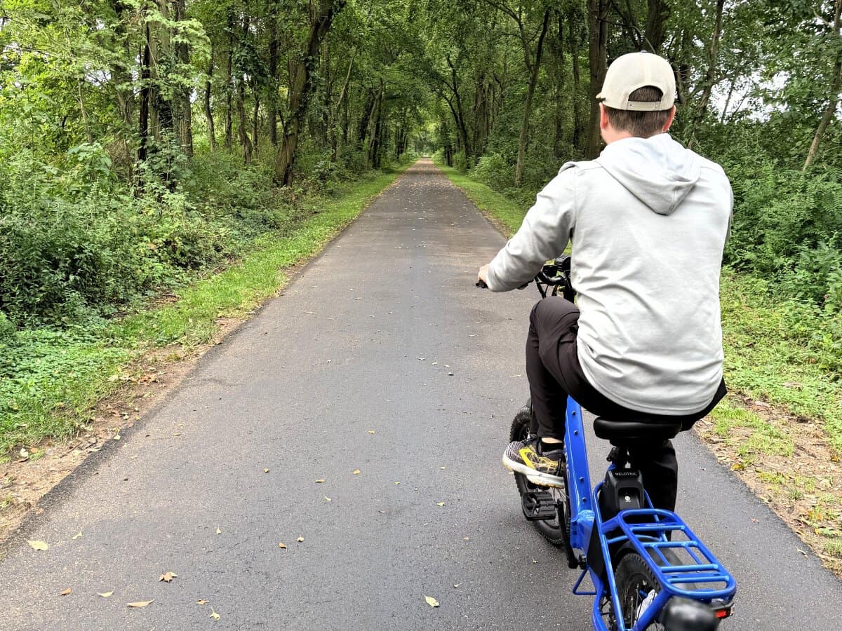 A cyclist on a blue e-bike rides away along a smooth, paved section of the Pumpkinvine Nature Trail beneath a canopy of green trees near Shipshewana, Indiana.
